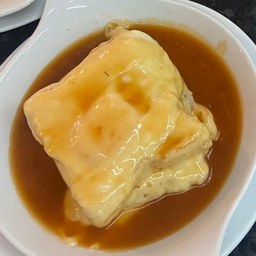Francesinha mit Ei, serviert in einer tiefen Schale, mit Sauce bedeckt